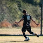 Young soccer player in black jersey with number 10 kicking a ball during a game on a sunny outdoor field.