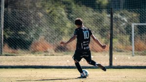 Young soccer player in black jersey with number 10 kicking a ball during a game on a sunny outdoor field.